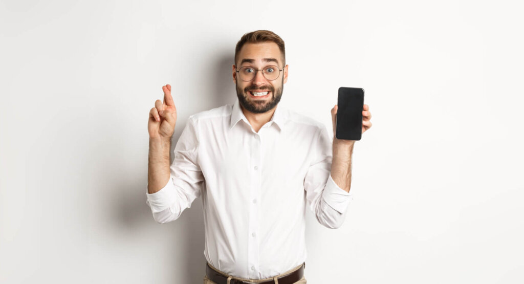 hopeful young business man showing mobile screen holding fingers crossed waiting online results scaled e1768592970797