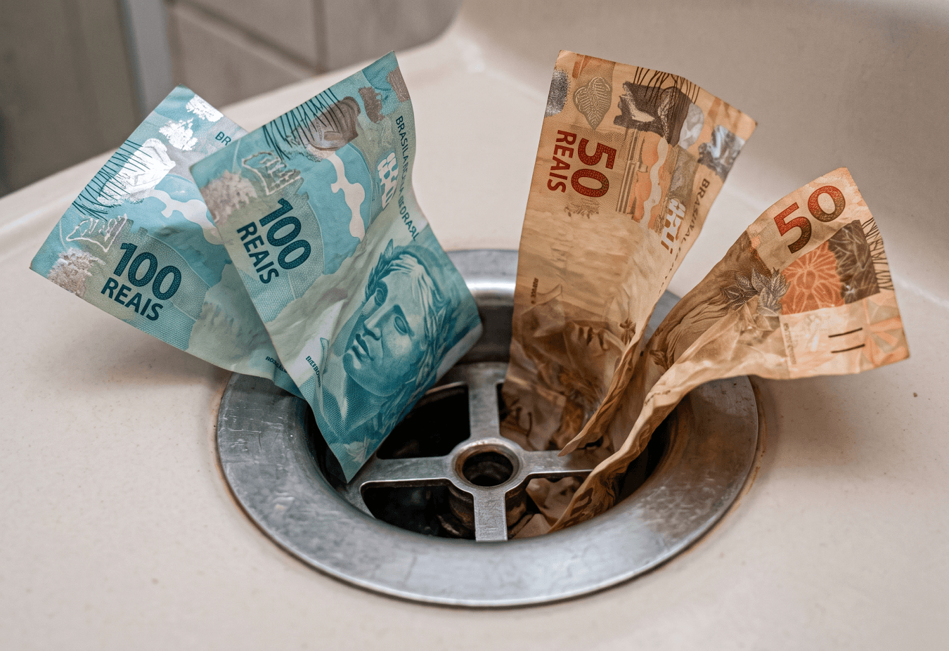 FindMe | Notas de Reais Brasileiros dentro de um ralo de pia, para representar a ideia de "Indo pelo ralo".